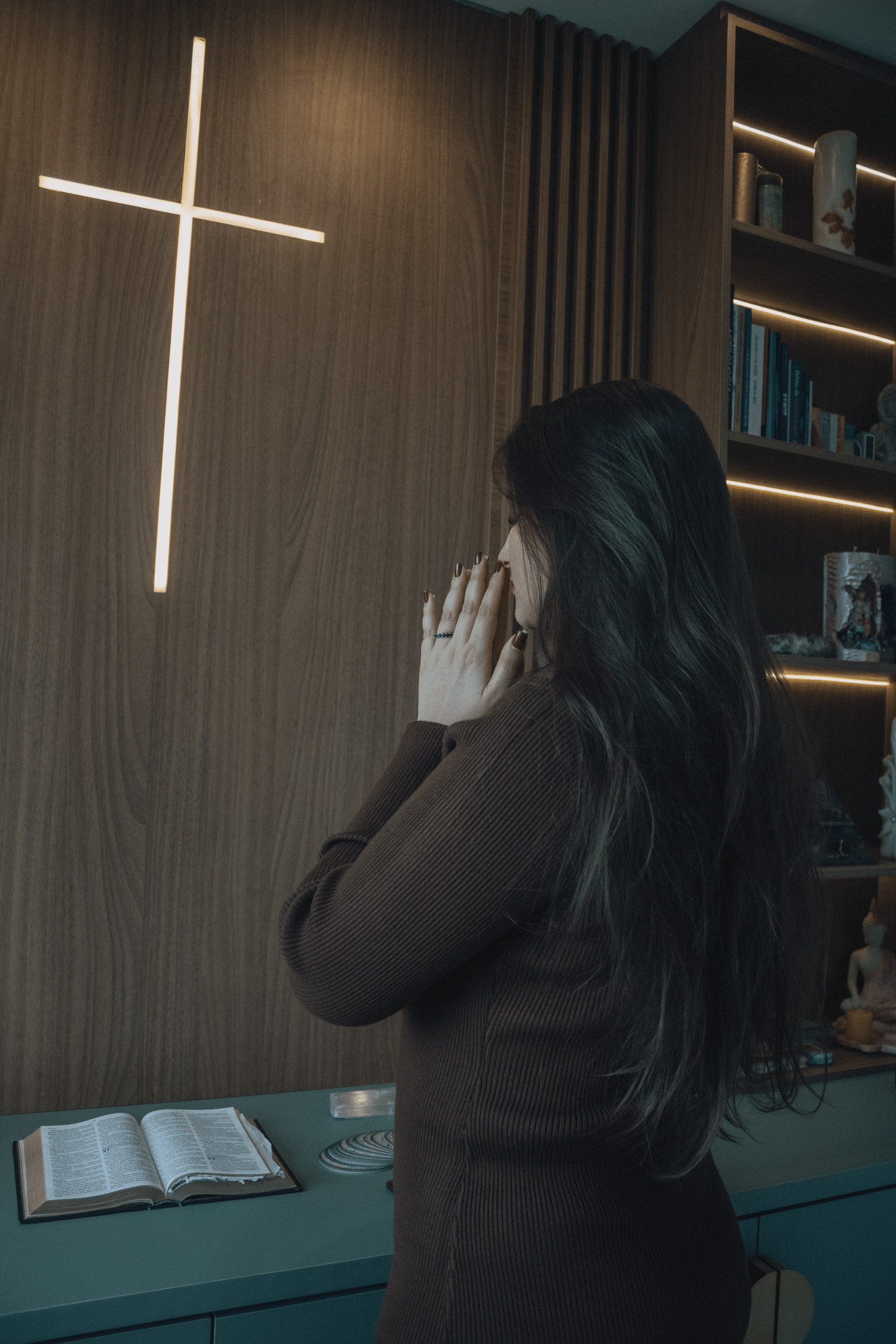 A woman prays in a serene room with an illuminated cross and open Bible.