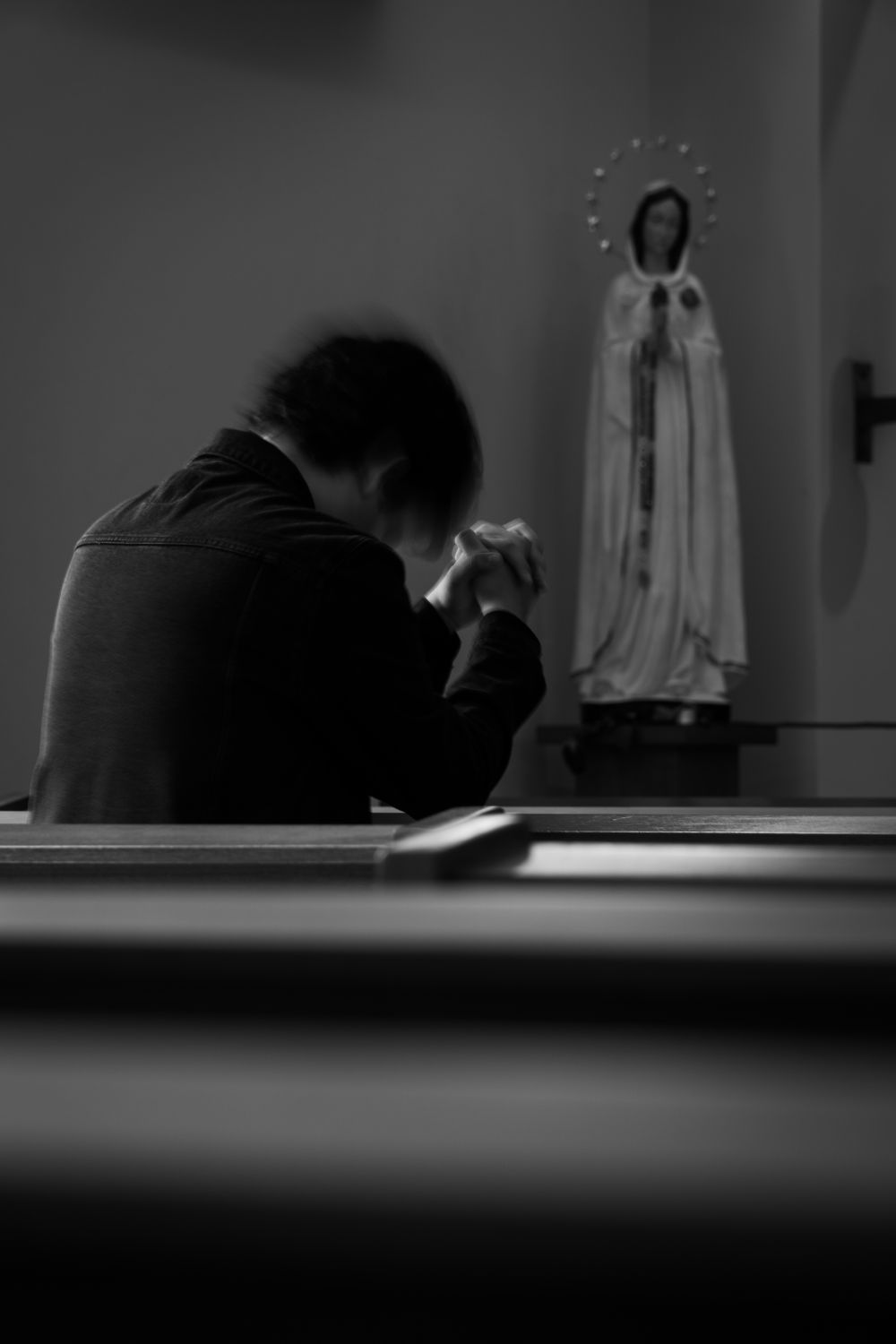 Black and white photo of a man praying beside a Virgin Mary statue in a church.