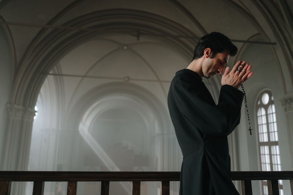 Side view of a priest praying with a rosary in a historic church setting.