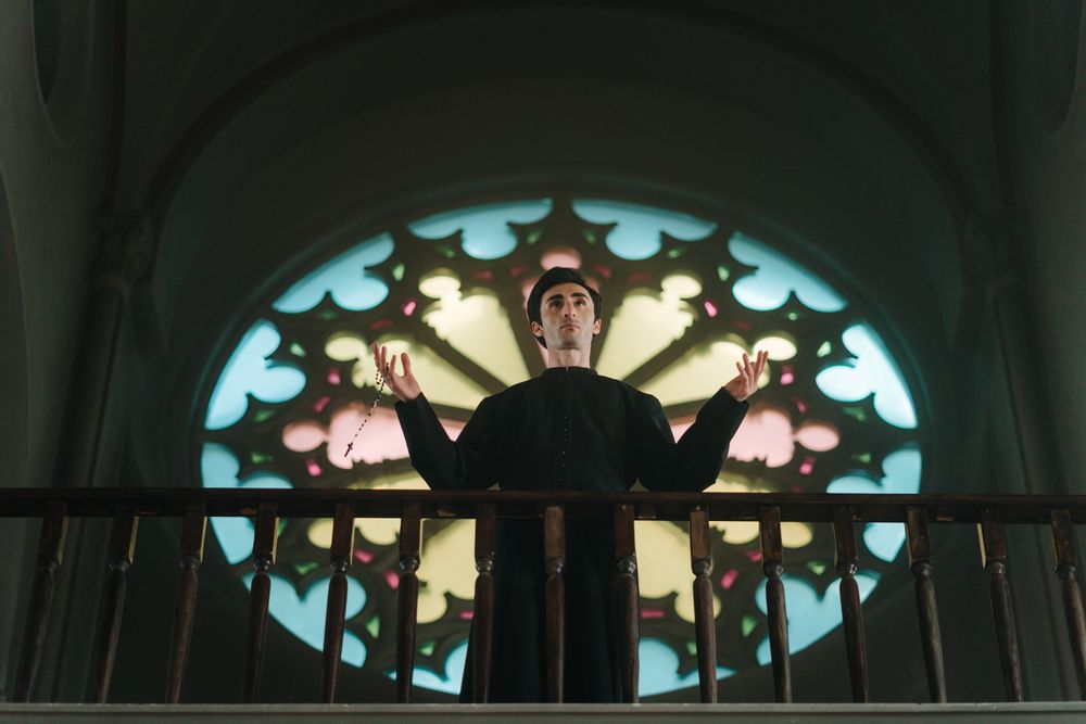 A priest stands in prayer holding a rosary in a church with stained glass.